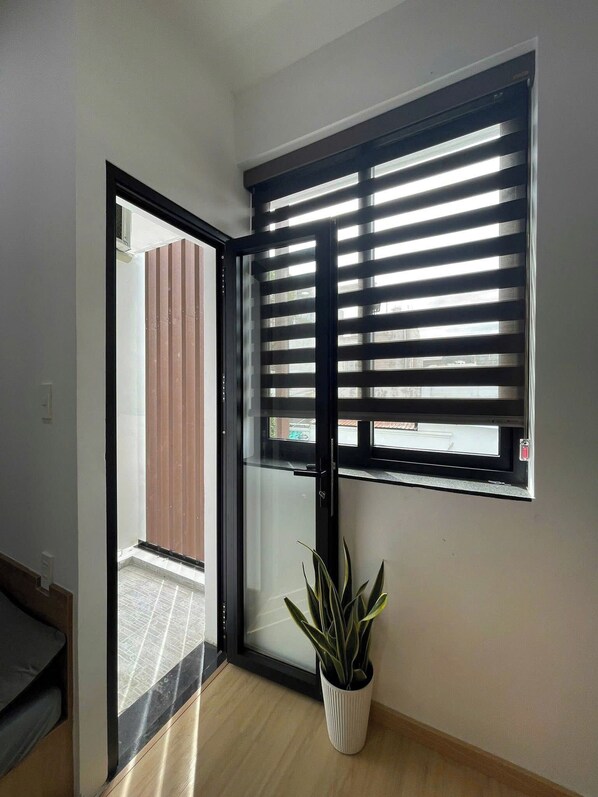 A room with a sliding glass door leading to an outdoor area, a window with horizontal blinds, and a potted plant on the floor. - Hat Nang Garden - Homestay & Restaurant (Ho Chi Minh City)