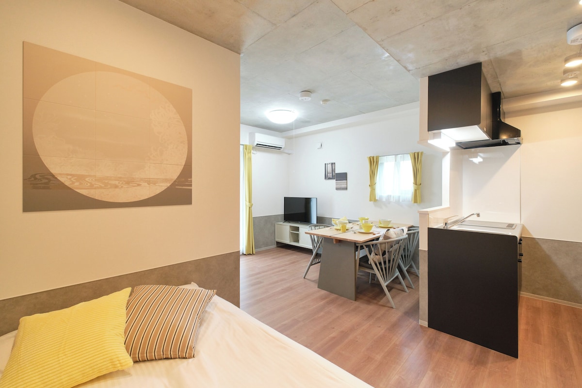 A modern hotel room with a bed, a dining area, and a kitchenette.