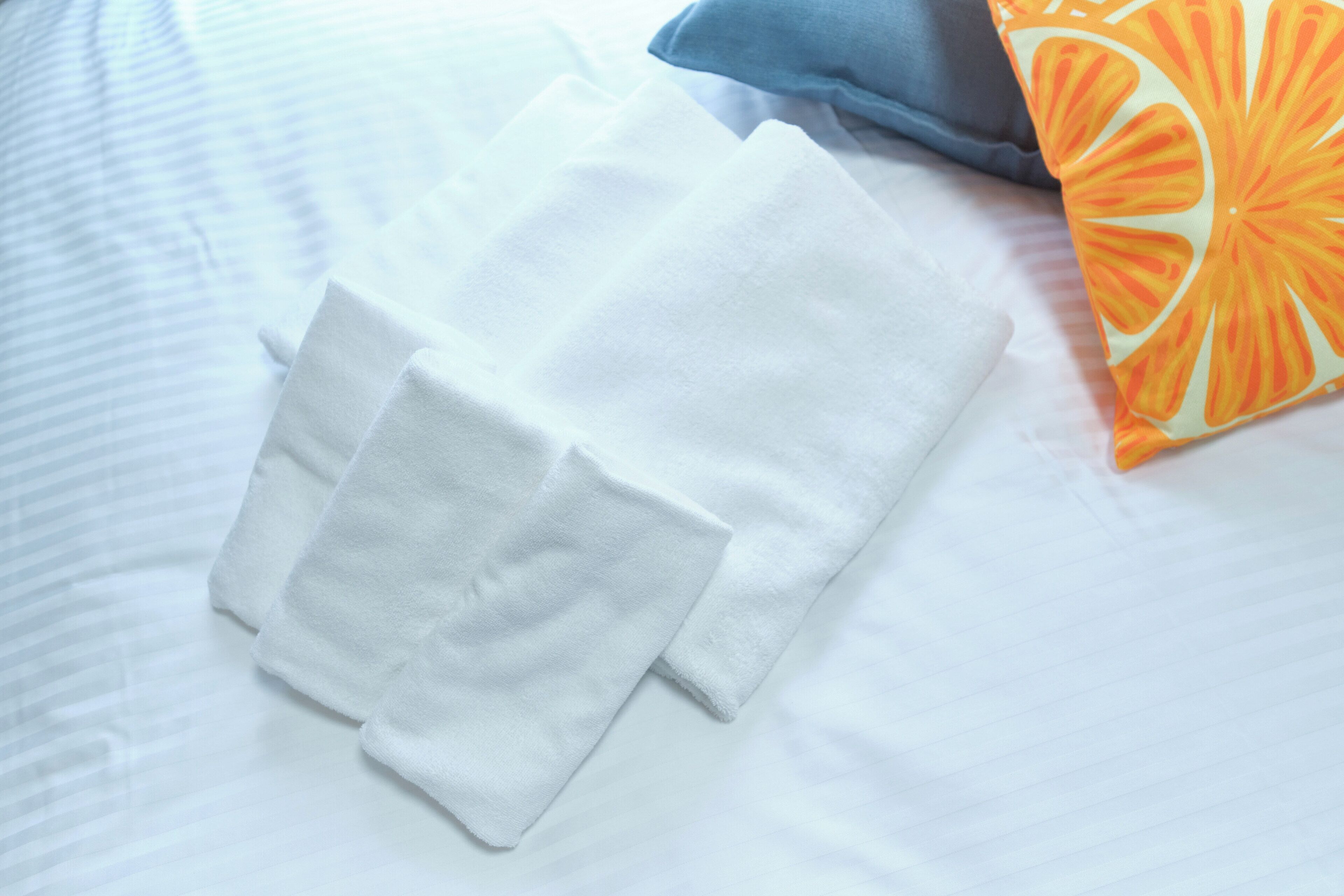 Neatly folded white towels on a bed with a striped blanket and a pillow with an orange design.