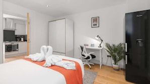 Habitación de hotel con cama, escritorio, silla, refrigerador, planta en maceta y kitchenette.