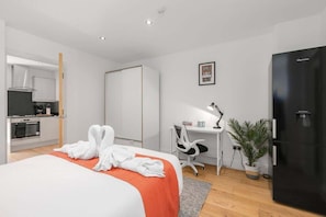 Ein Hotelzimmer mit Bett, Schreibtisch, Stuhl, Kühlschrank, einer Topfpflanze und einer Küchenzeile.
