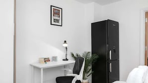 Una habitación pequeña y moderna con un escritorio blanco, una silla negra, un refrigerador negro y un cuadro en la pared.