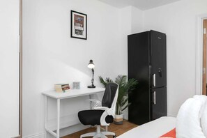 Ein kleiner, moderner Raum mit einem weißen Schreibtisch, einem schwarzen Stuhl, einem schwarzen Kühlschrank und einem gerahmten Bild an der Wand.