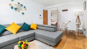Una sala de estar moderna con un sofá seccional gris, un comedor con una mesa y sillas blancas, y una pequeña cocina visible a través de una puerta abierta.
