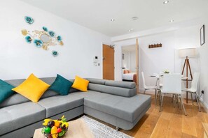 Ein modernes Wohnzimmer mit einem grauen Ecksofa, einem Essbereich mit einem weißen Tisch und Stühlen und einer kleinen, durchgehende Küche.