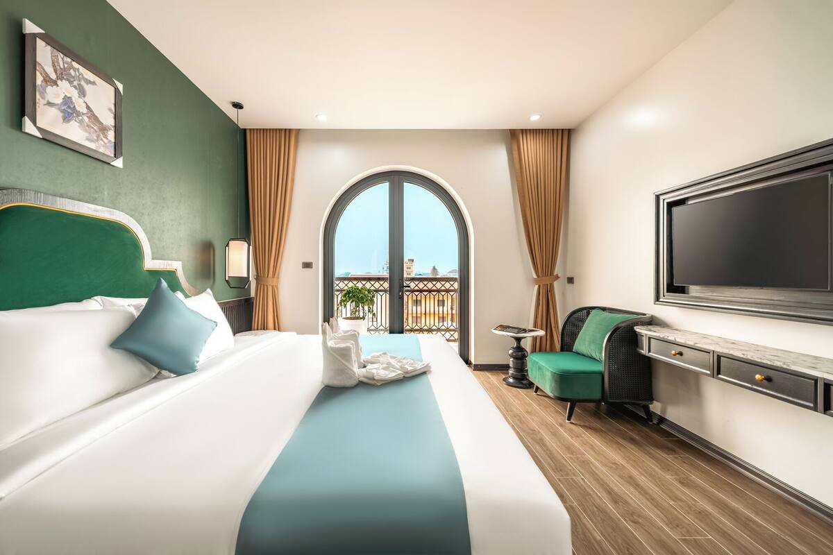 A hotel room with a bed, a desk, a TV, a balcony with a view, and a painting on the wall.
