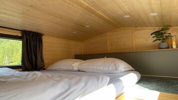 A wooden cabin room with a bed, a window with curtains, a plant on a shelf, and a candle.