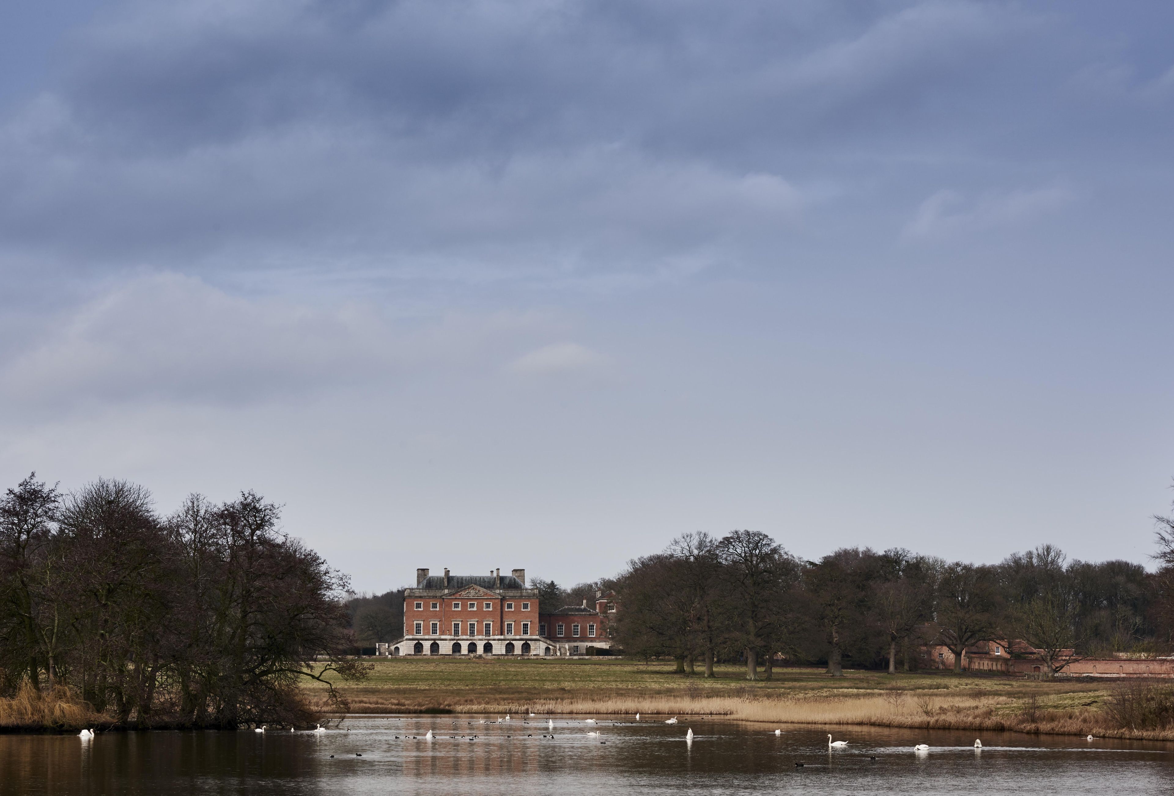 A large brick building with a symmetrical facade and a central entrance, surrounded by a body of water with swans and ducks.