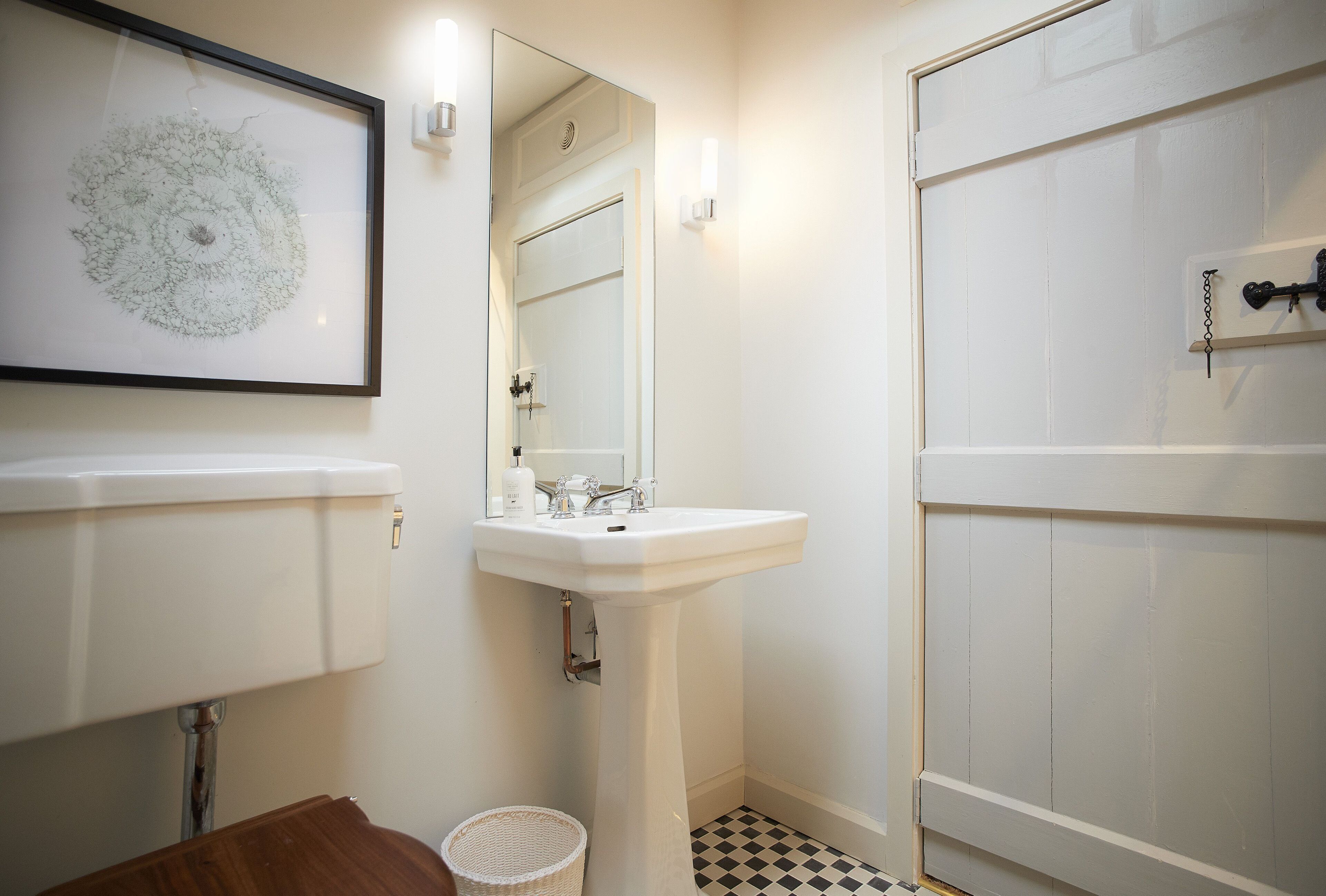 Une salle de bain avec un lavabo blanc, un miroir et des toilettes. Un tableau encadré est accroché au mur et un petit panier est posé sur le sol.