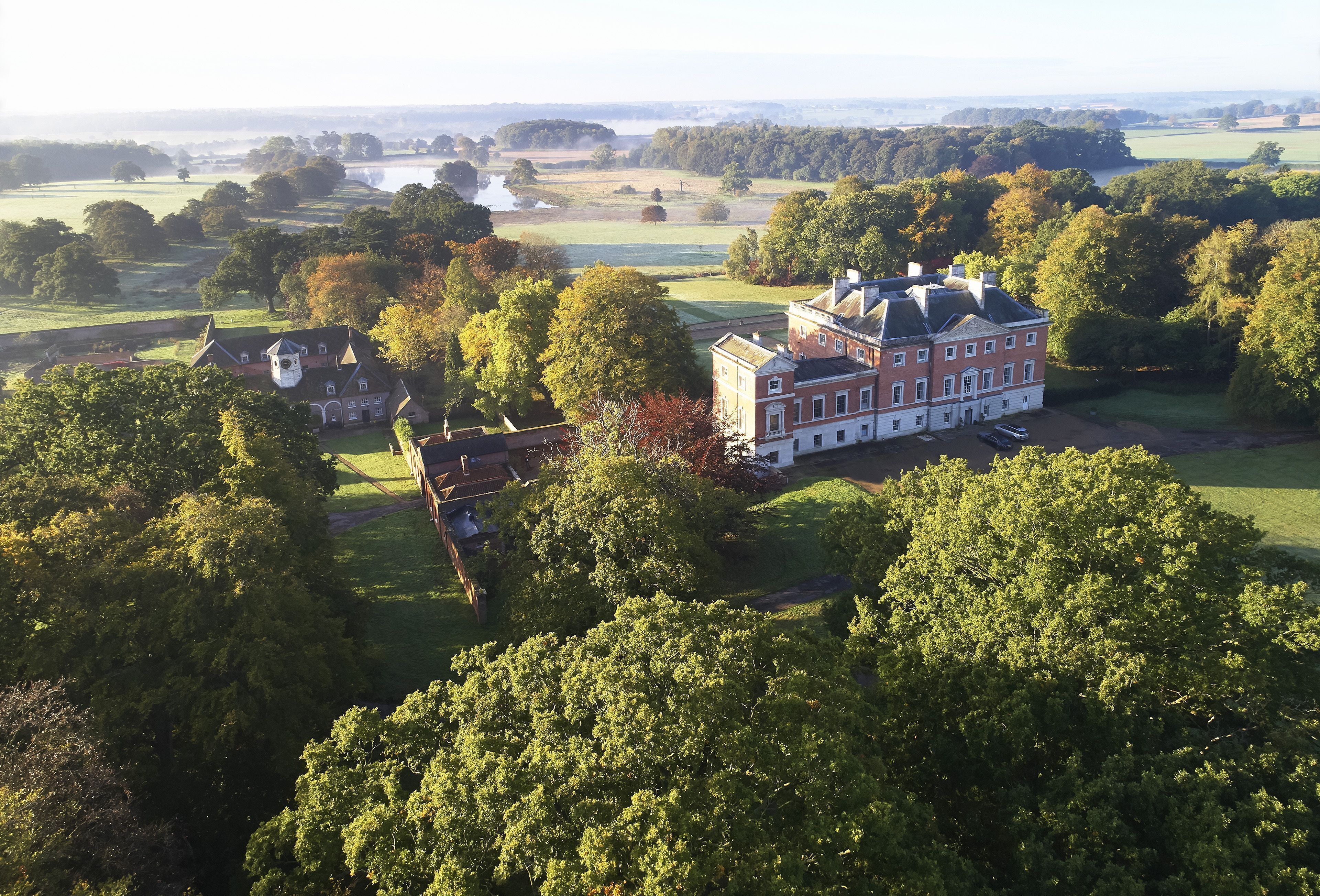A large estate with multiple buildings, including a prominent red brick house with a white tower, surrounded by lush greenery and open fields.