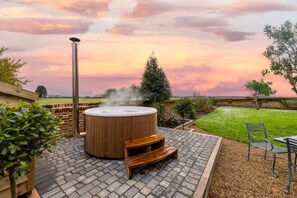 A hot tub in a garden with a brick patio, surrounded by greenery and a clear sky.