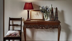 Una mesa de madera con tapa tallada, una lámpara roja, un cuadro enmarco y un jarrón con flores.