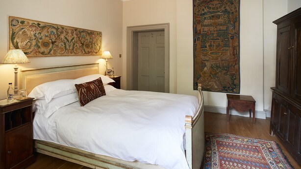A bedroom with a large bed, a nightstand with a lamp, a wooden dresser, and a tapestry on the wall.