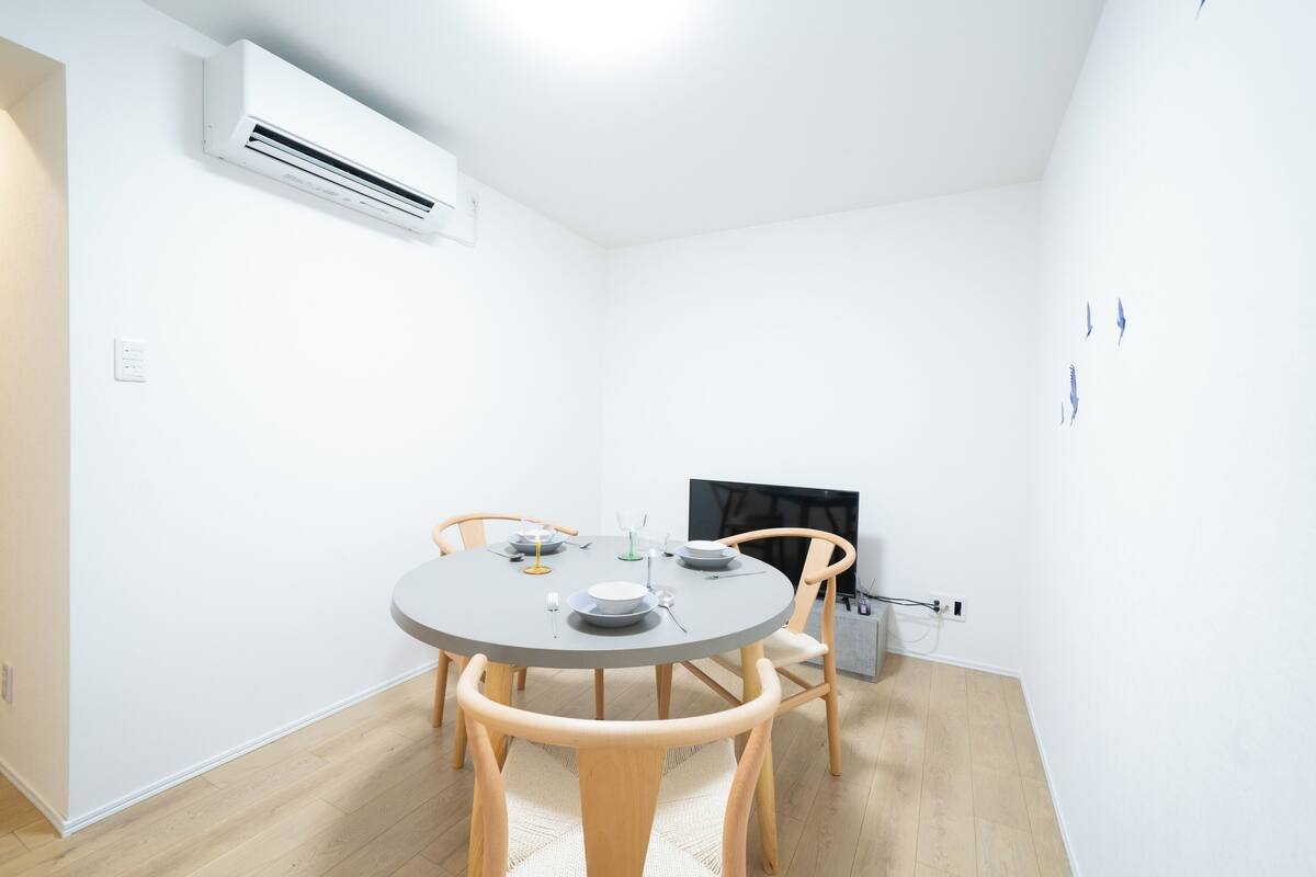 A minimalist room with a round table, four wooden chairs, a flat-screen TV, and an air conditioning unit mounted on the wall.
