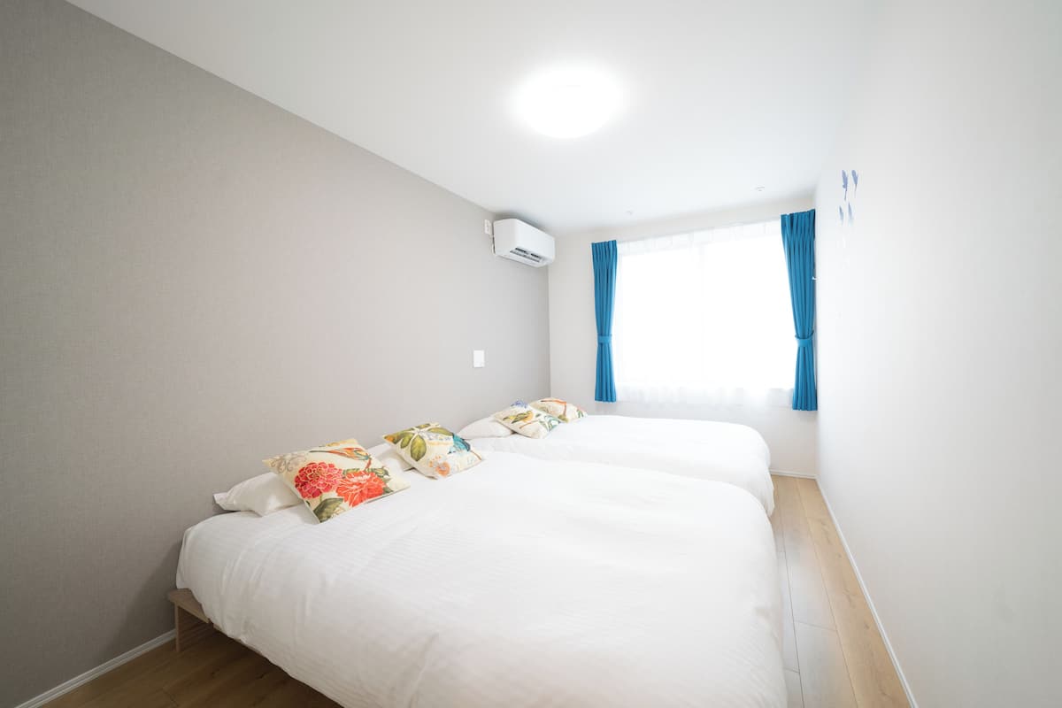 A room with two beds, a window with curtains, an air conditioning unit, and a wooden floor.