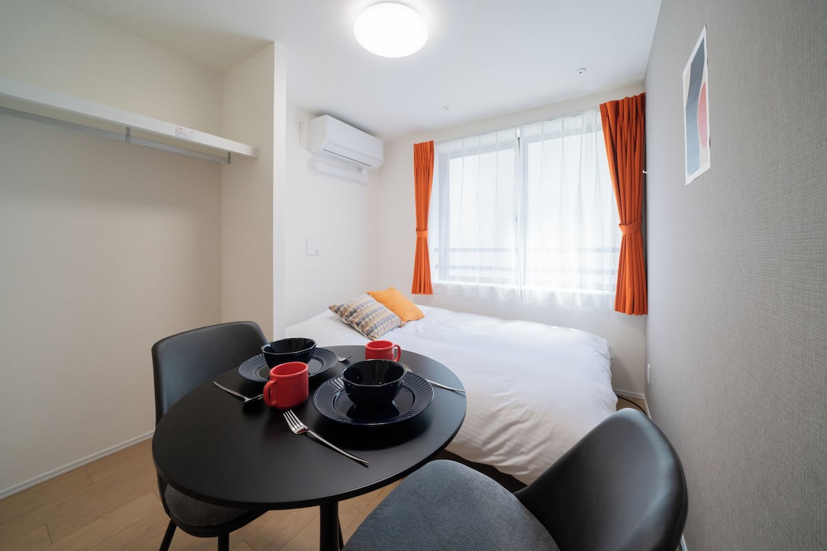 A hotel room with a bed, a table with red cups and plates, and a window with orange curtains.