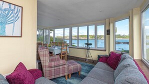 Une véranda avec vue sur la mer, un coin repas avec une table et des chaises, et un grand canapé gris agrémenté de coussins rouges.