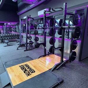 Un gimnasio con equipo de levantamiento de pesas, una plataforma de madera con la marca 'HAMMER STRENGTH' y un muro con espejos.