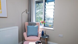 Una habitación acogedora con un sill ón rosa, una mesita de noche con un jarrón de flores, un radiador y una ventana con vistas a árboles.
