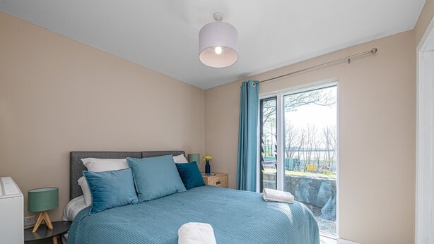 Un dormitorio con cama, mesita de noche, lámpara y vistas al exterior.