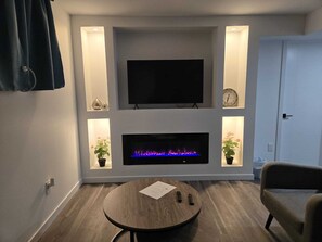 A modern living room with a flat-screen TV mounted above a fireplace, flanked by two small shelves with decorative items. - Cozy private bsmt suite |Bonavista |Fireplace |Workspace & nature-inspired decor (Winnipeg)