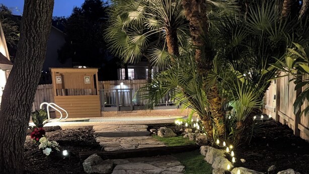 A stone pathway with lights, surrounded by rocks and plants, leading to a wooden deck. - Cozy Luxury  Pool + Home Theater + Backyard Oasis (San Antonio)