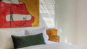 Un letto con biancheria bianca e un cuscino verde, una testiera in legno, un quadro appeso al muro e un tavolino di vimini con una lampada.