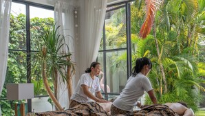 Una stanza con due lettini da massaggio, un'ampia finestra con tende e vista su piante tropicali.