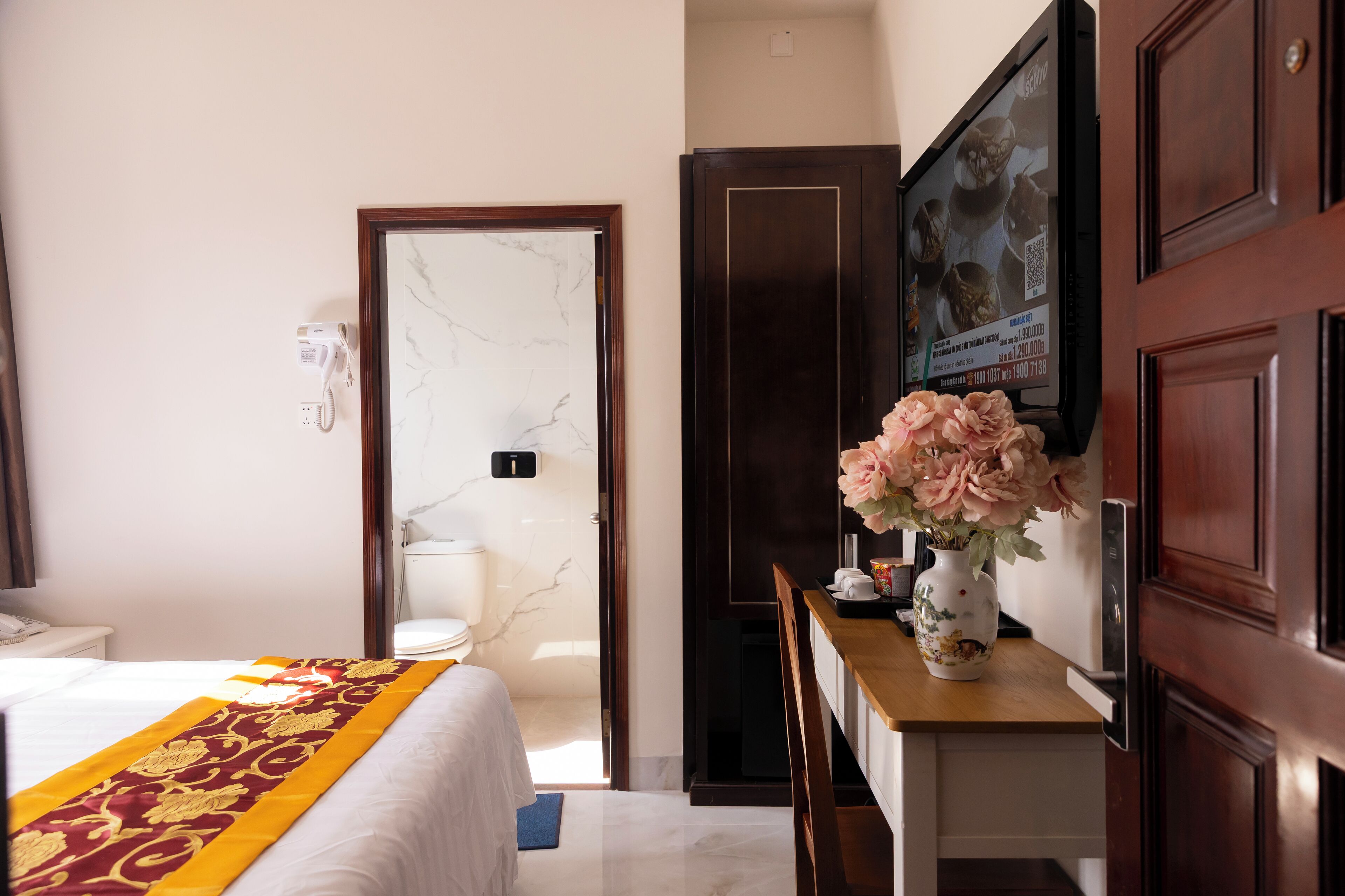 A hotel room with a bed, a desk with a vase of flowers, a television, and a door leading to the bathroom.