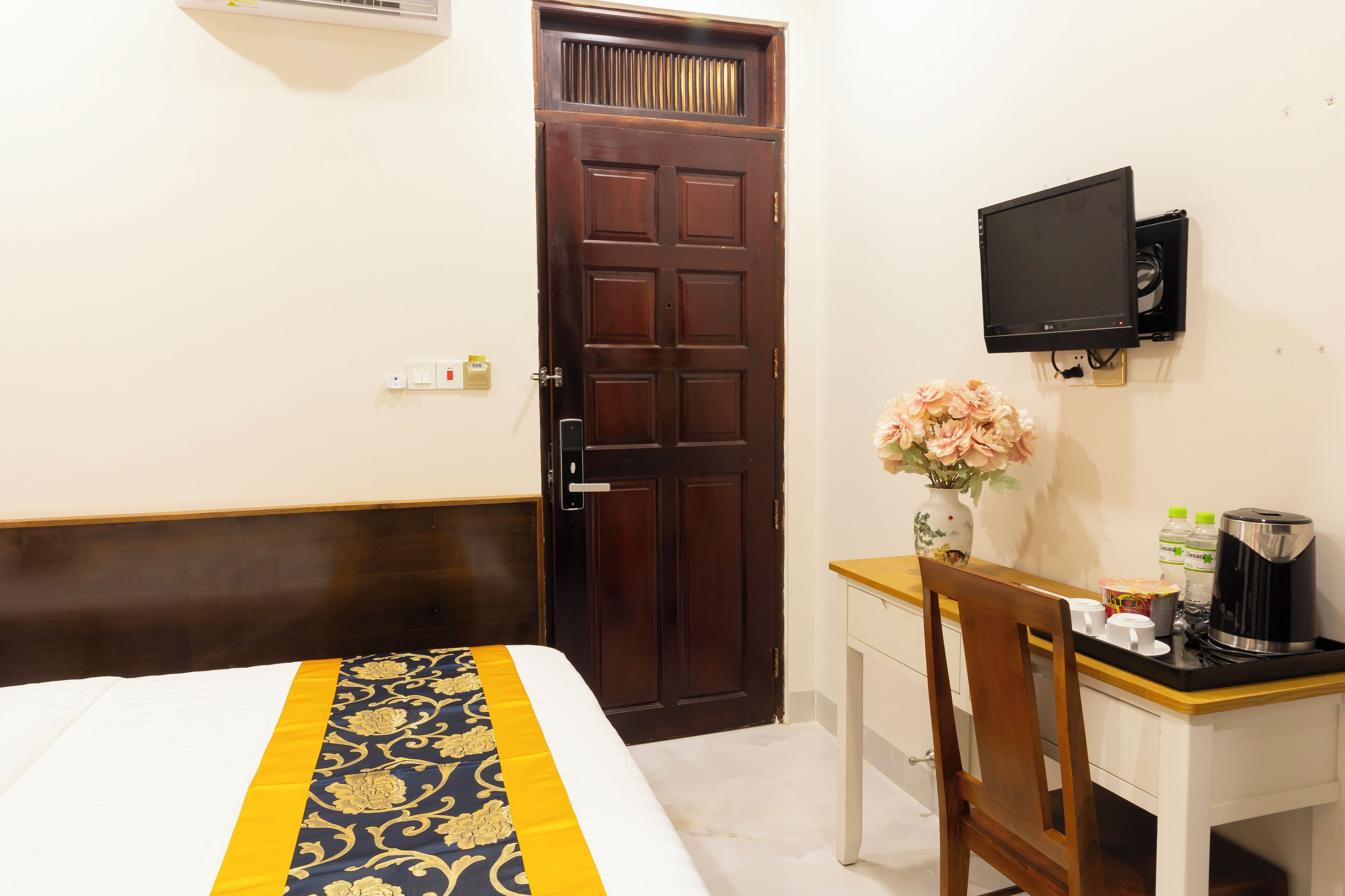 A hotel room with a bed, a desk, a TV, a vase with flowers, and a coffee maker.