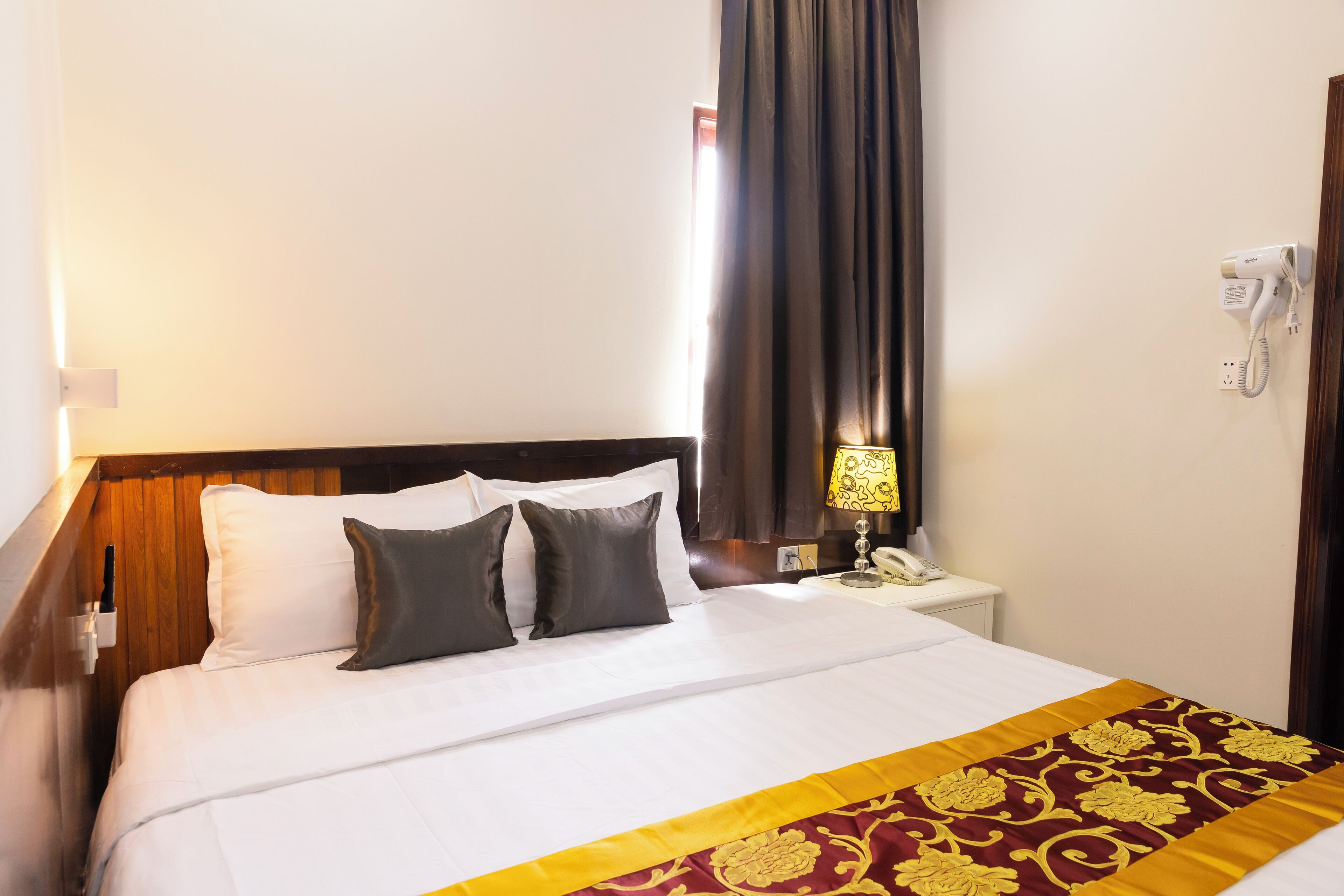 A hotel room with a neatly made bed, a bedside lamp, a telephone, and a window with curtains.
