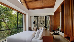 A modern bedroom with a large bed, a desk, and a view of trees through large windows. - Uma Santai Villa Canggu by Nakula (Canggu)