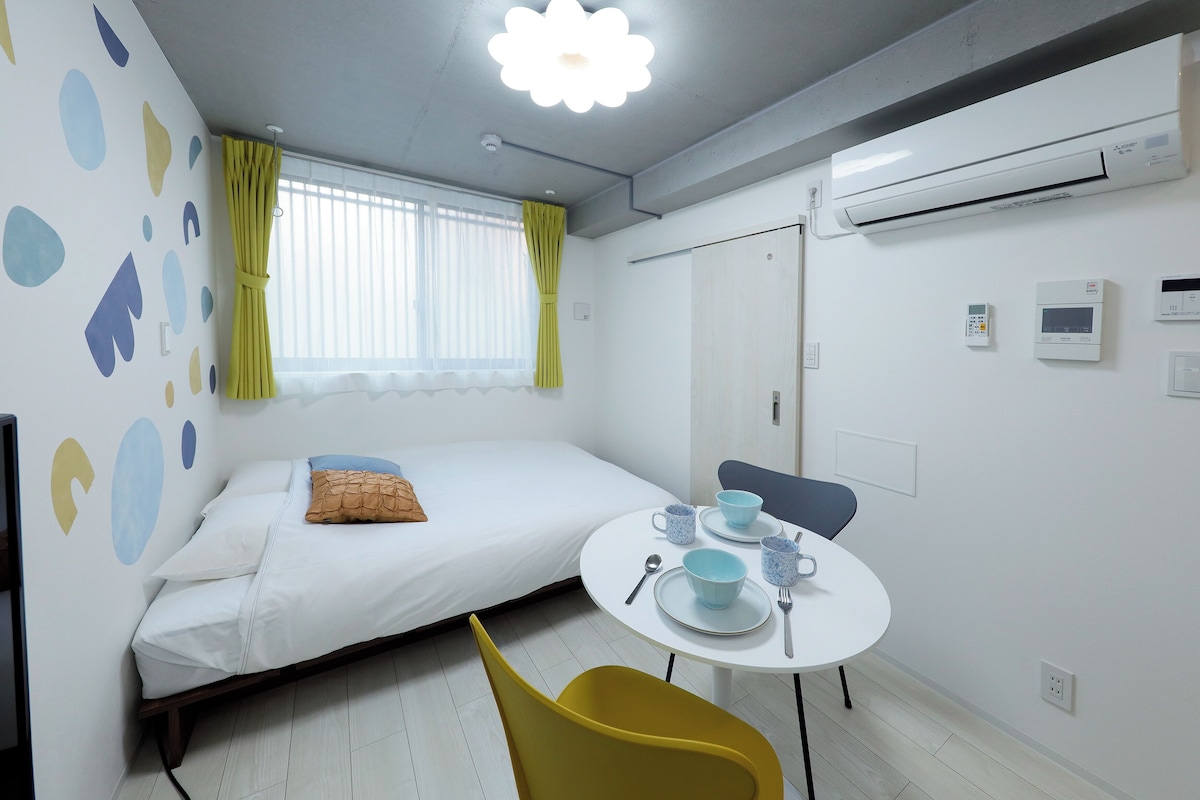 A small, modern room with a bed, a round table with a chair, and a window with curtains.