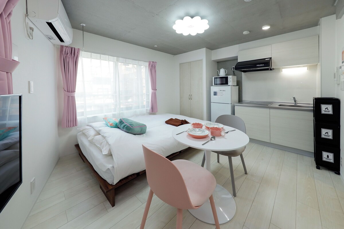 A compact hotel room with a bed, a dining table with chairs, a kitchenette, and a large window with curtains.