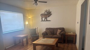 A living room with a wooden floor, a sofa, a coffee table, a chair, a lamp, and a wall-mounted deer decoration.