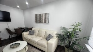 A living room with a beige leather sofa, a coffee table, a potted plant, and a flat-screen TV mounted on the wall. - Three Bedroom Bungalow with free parking, closer to commercial area and highway (Mississauga)