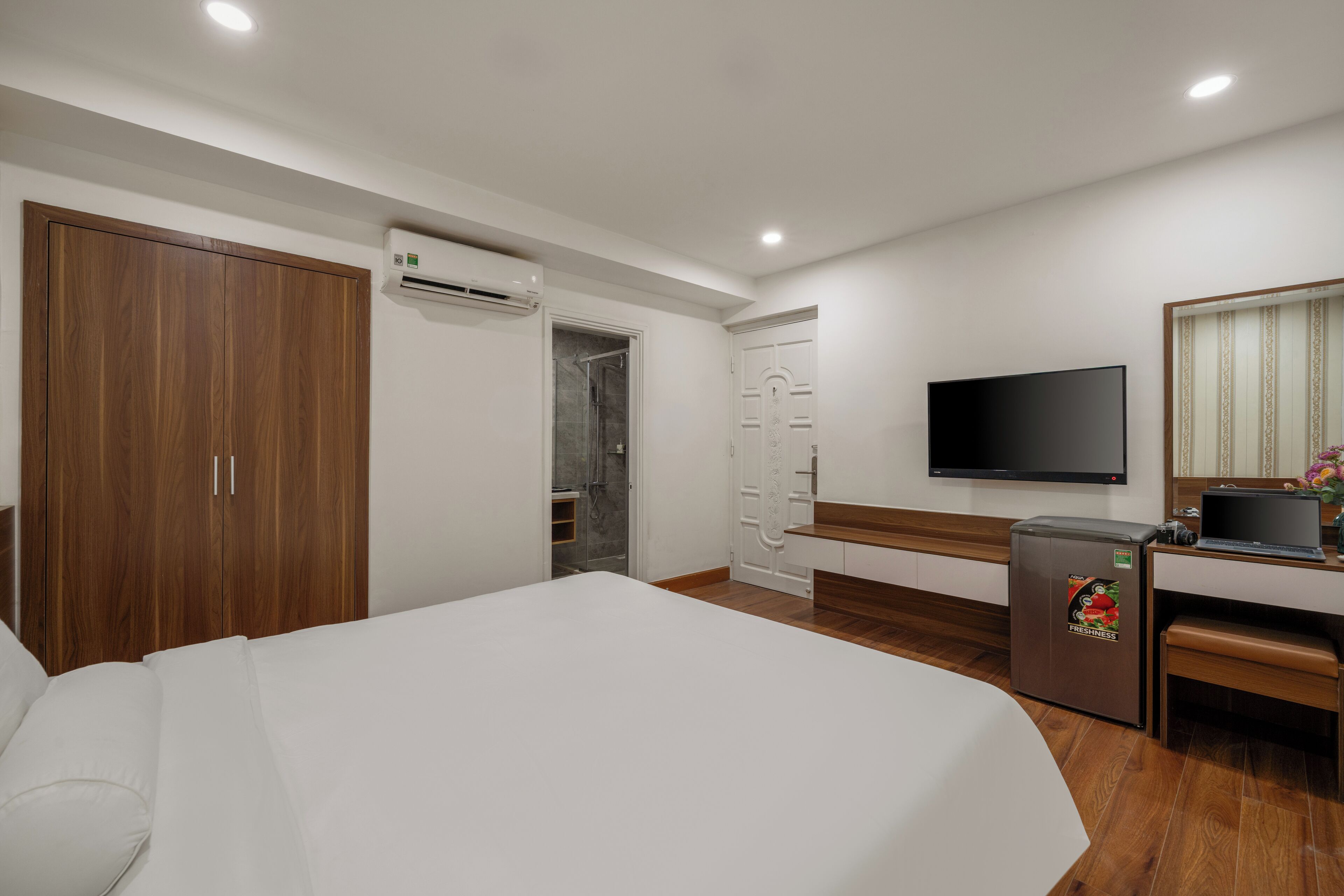 A hotel room with a large bed, a wooden wardrobe, an air conditioning unit, a shower, a television, and a small refrigerator.
