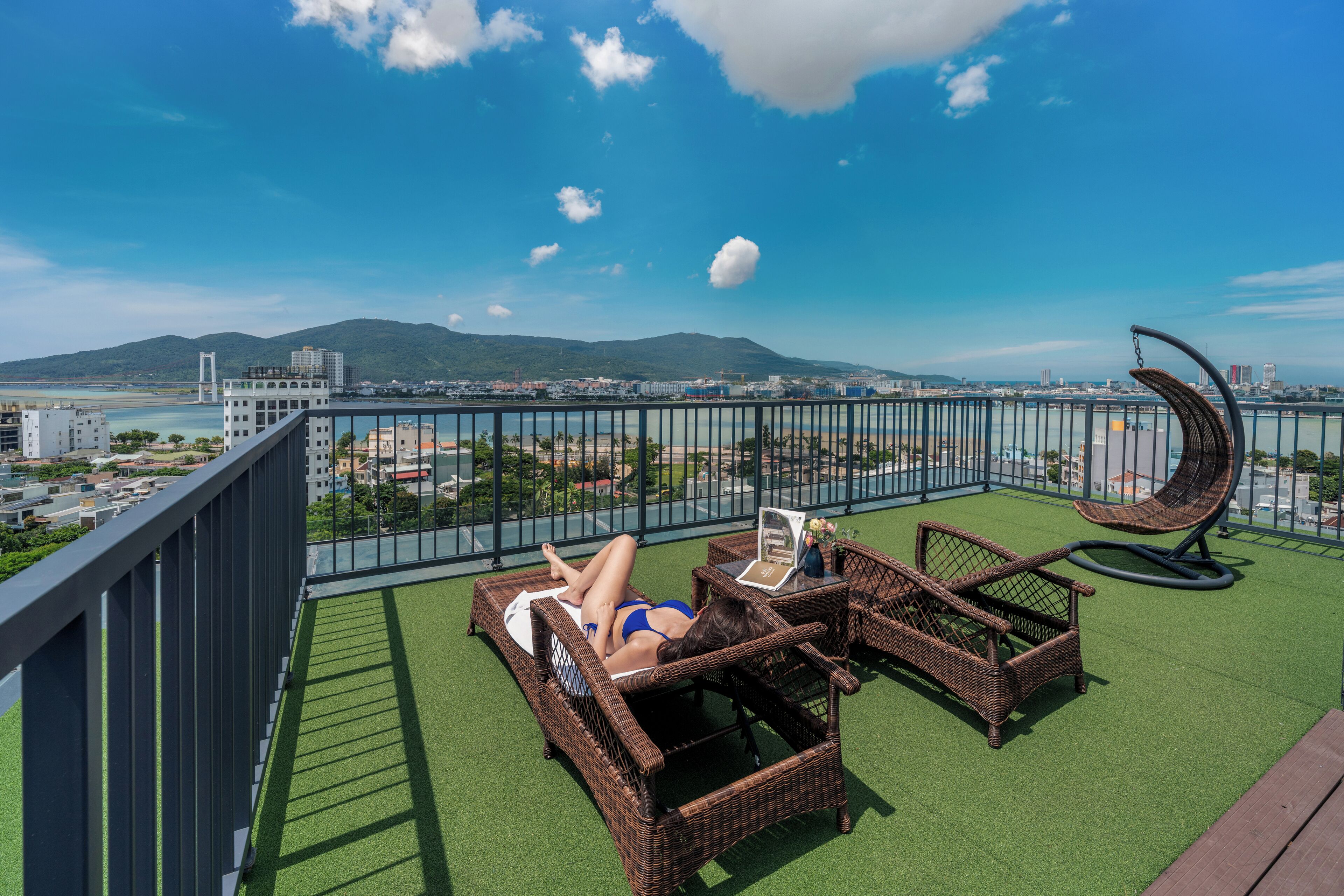 A balcony with a view of a cityscape, including a bridge and mountains. There are wicker lounge chairs and a small table with a chair.