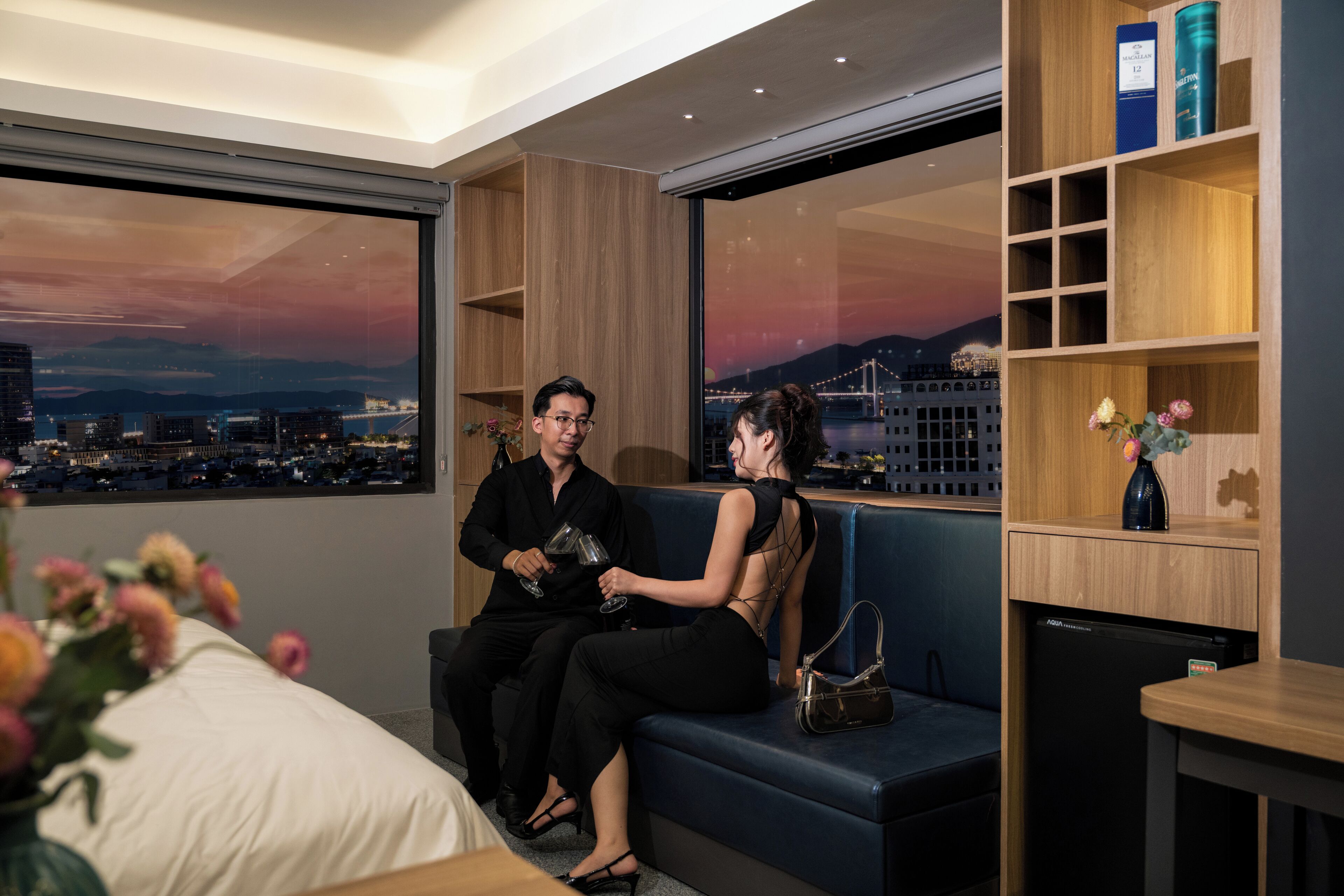 A modern hotel room with a city view, a sofa, a small table, and a bookshelf.