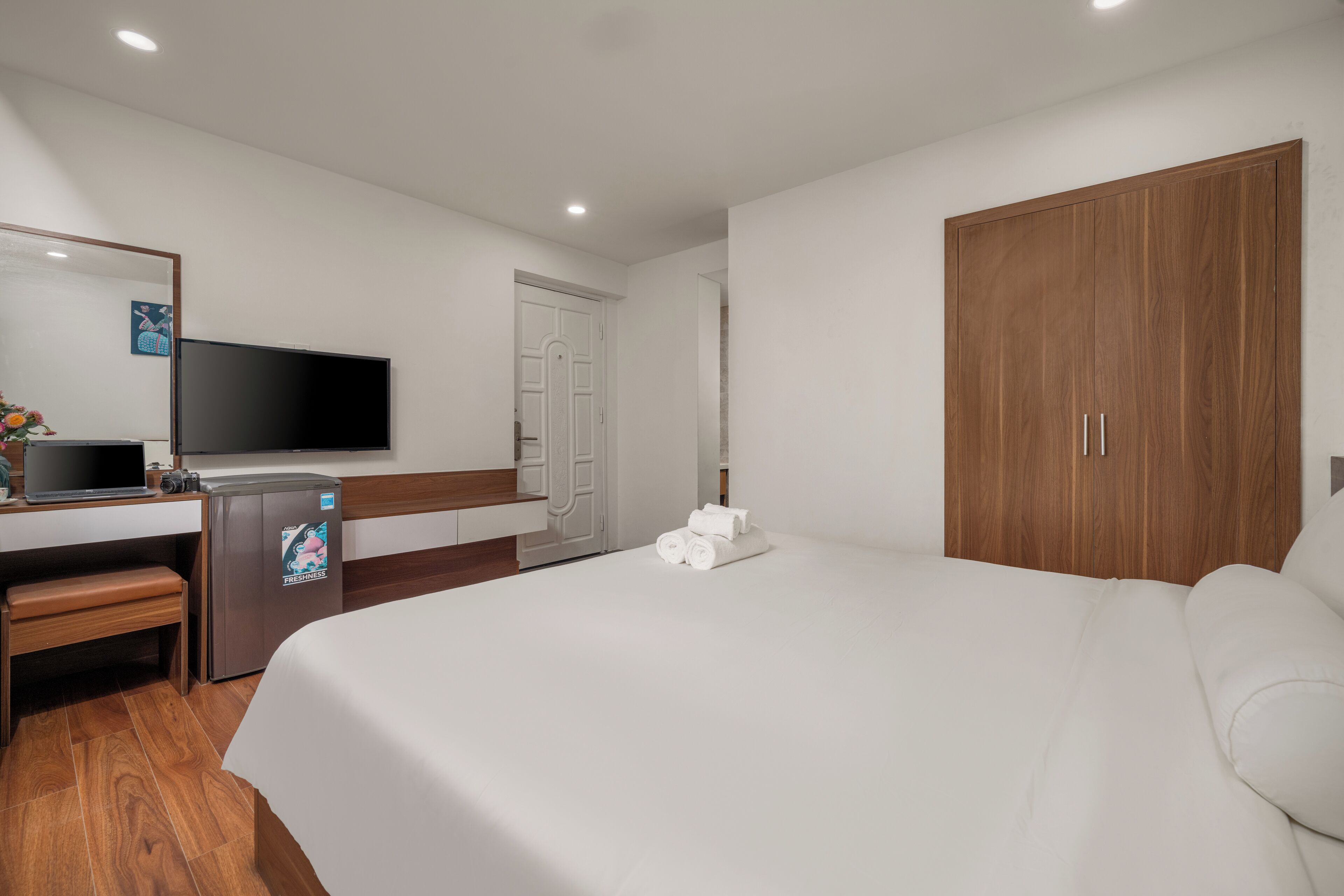 A hotel room with a bed, a desk with a computer and a refrigerator, a television, and a wardrobe.