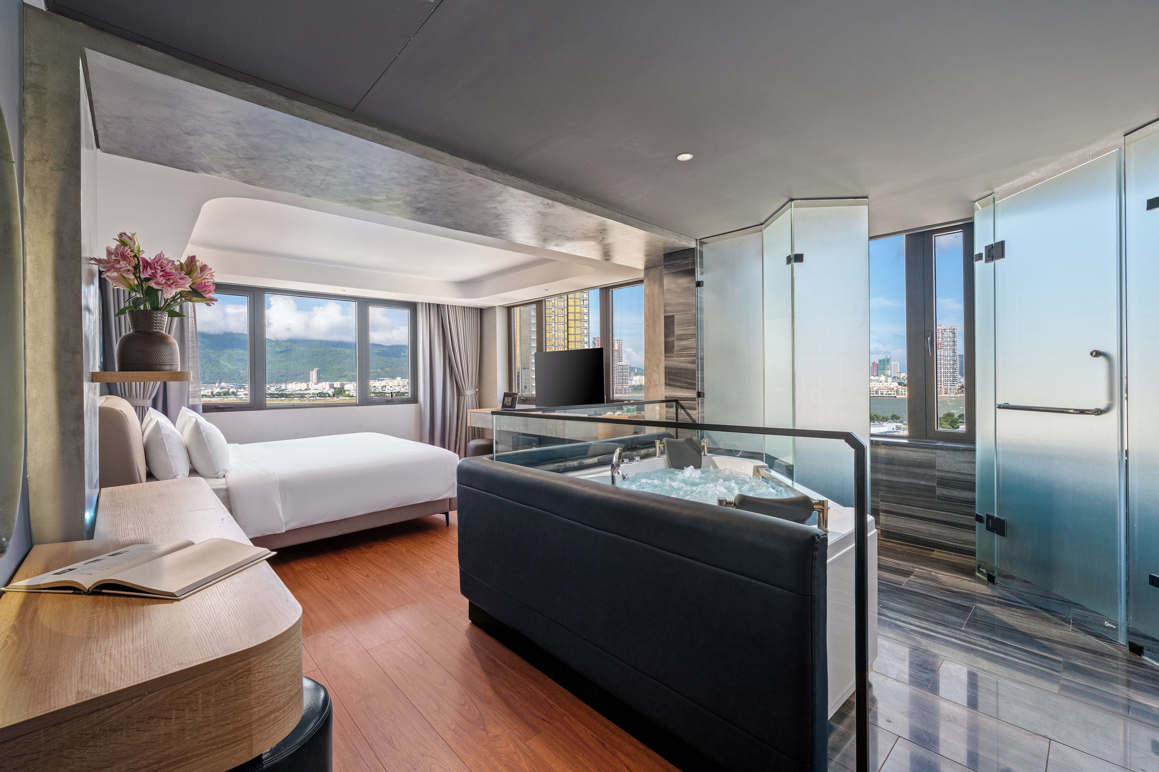 A modern hotel room with a bed, a seating area, a glass-enclosed jacuzzi, and a city view.