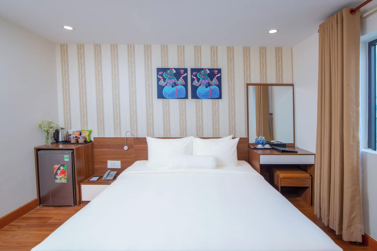 A hotel room with a large bed, a desk, a small refrigerator, a mirror, and a window with a view of buildings and water.