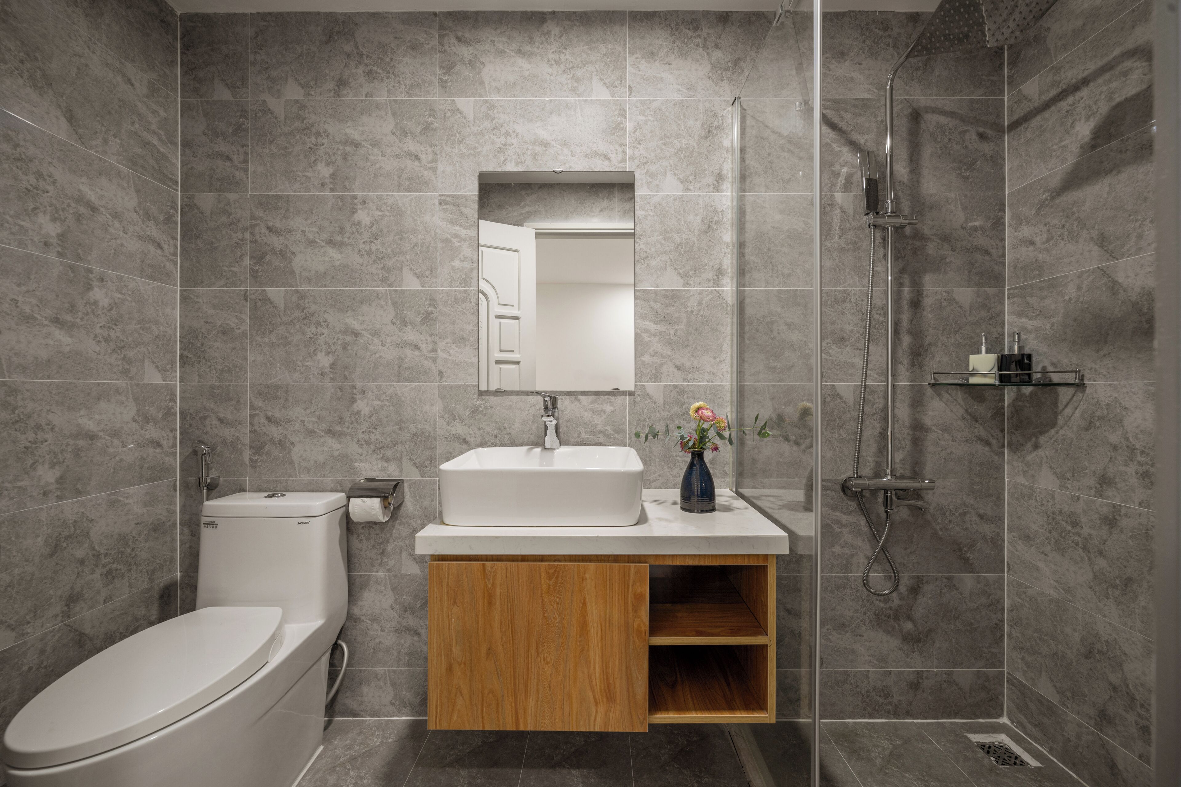 A modern bathroom with a toilet, sink, mirror, shower, and wooden cabinet.
