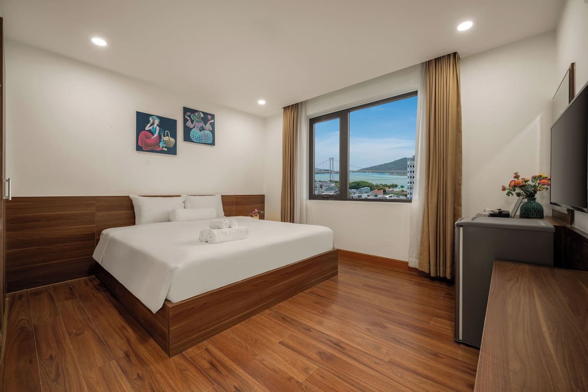 A hotel room with a large bed, wooden flooring, a desk, a TV, and a window with a city view.