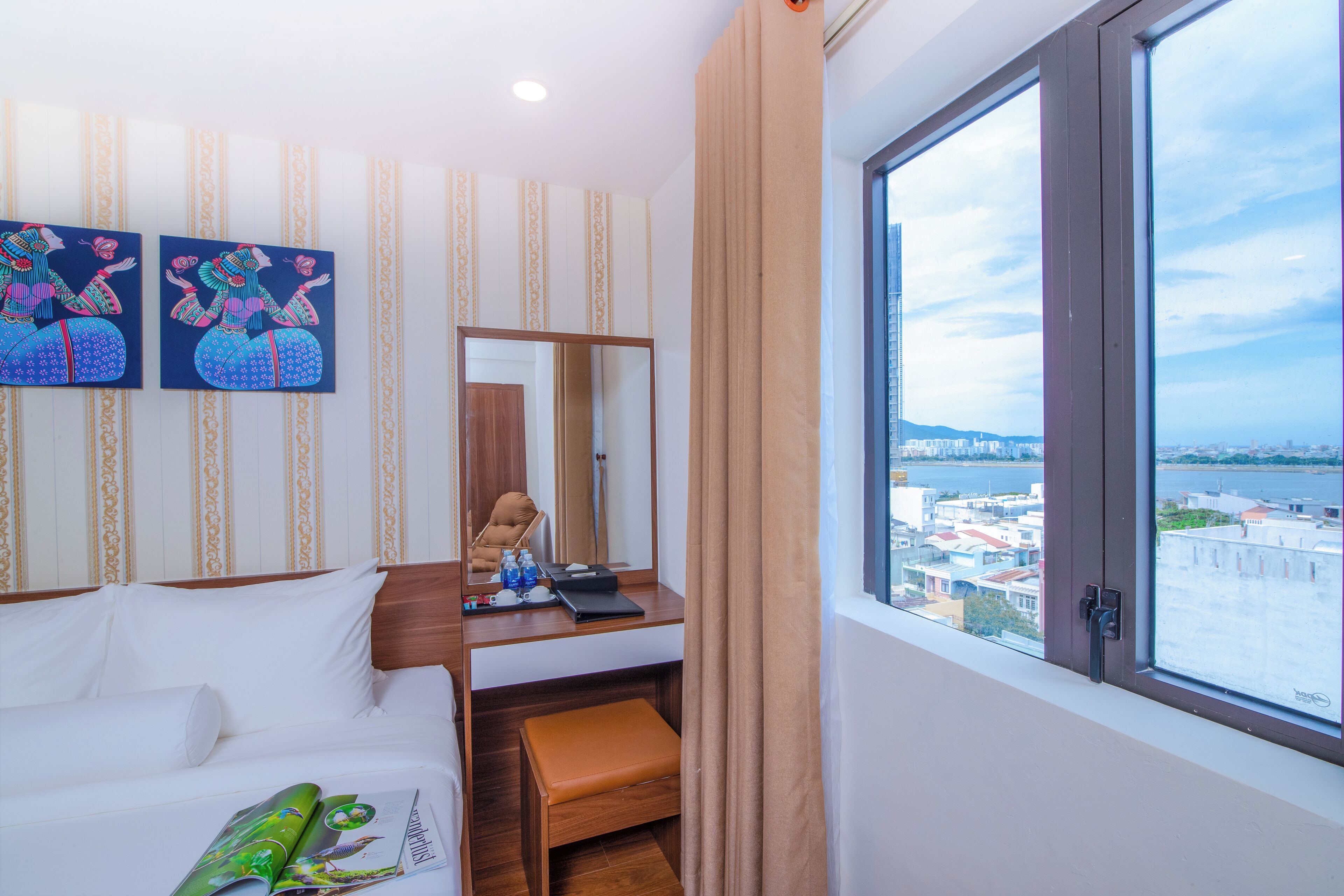 A hotel room with a bed, a desk with a laptop, a mirror, and a view of the city through a large window.