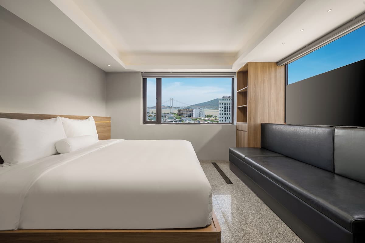 A modern hotel room with a large bed, a black leather sofa, a TV, and a window with a city view.