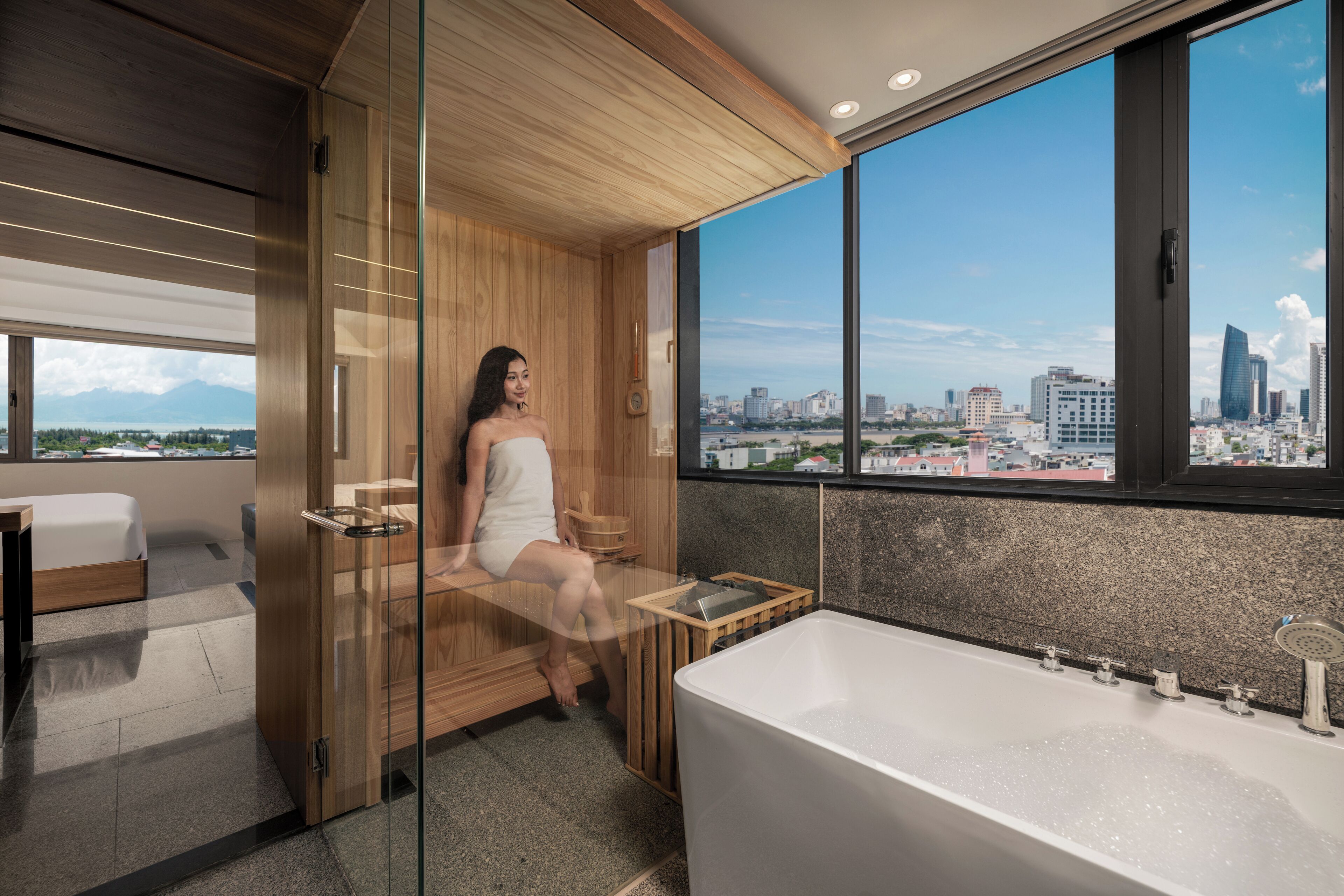 A modern bathroom with a large bathtub, a wooden sauna, and a city view through large windows.