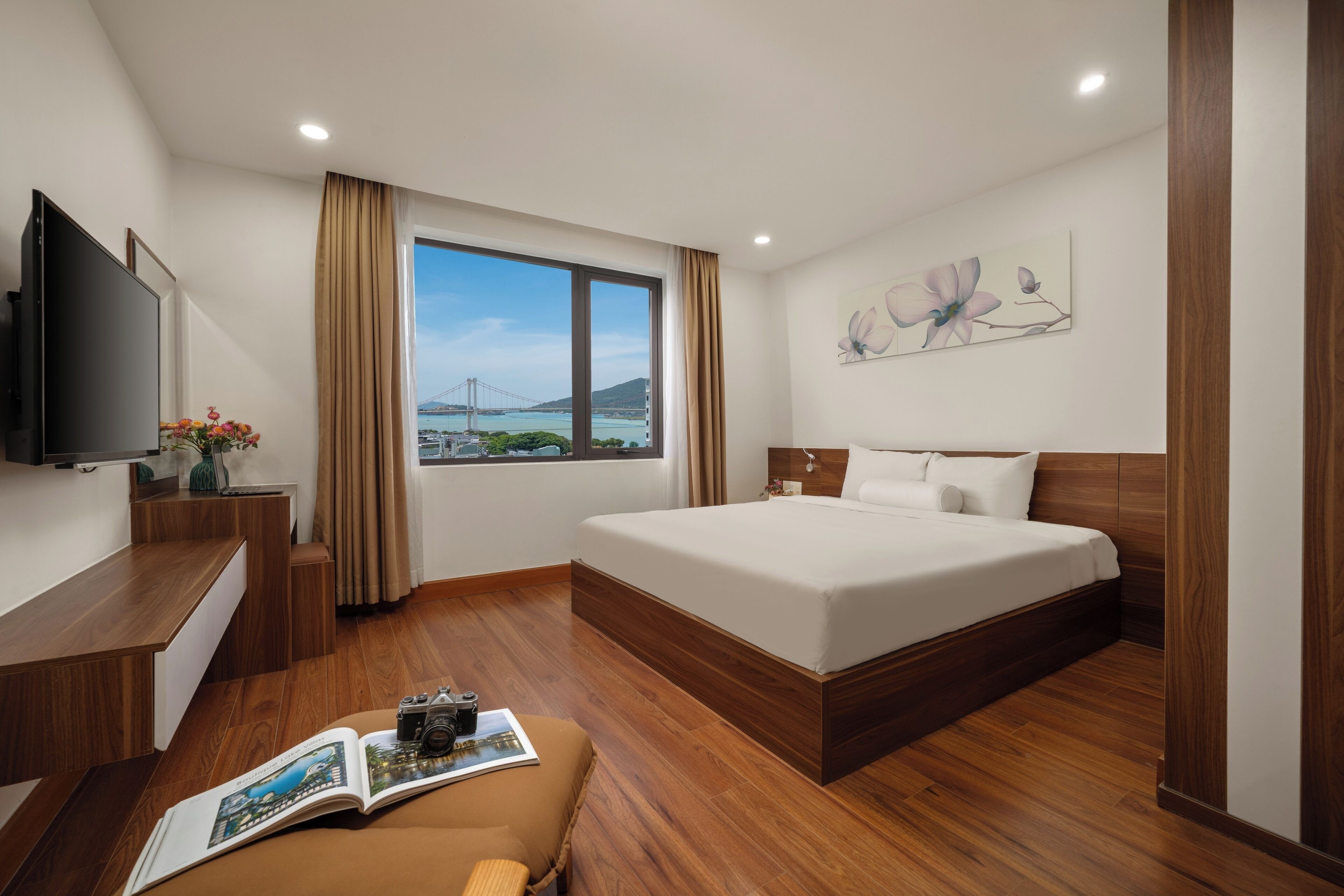 A modern hotel room with a large bed, wooden flooring, a TV, and a window with a view of a bridge and mountains.