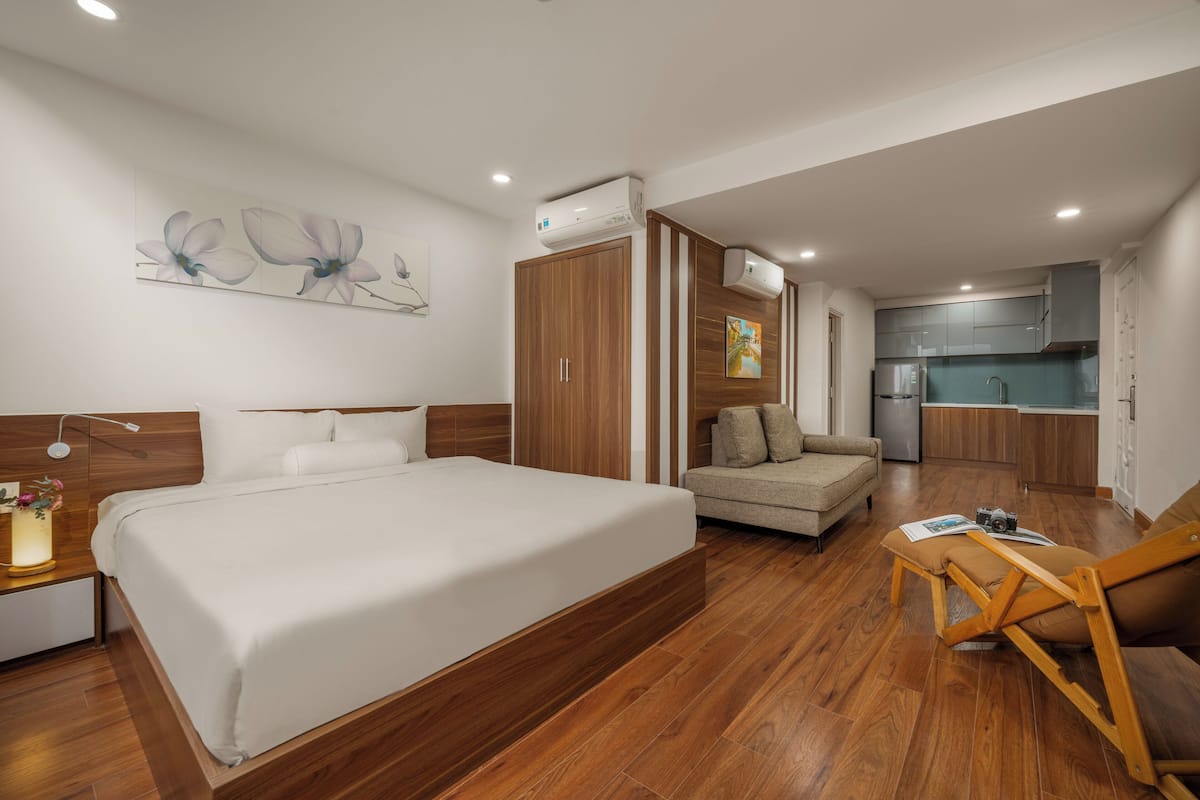 A modern hotel room with a large bed, a sofa, a wooden floor, and a kitchenette in the background.