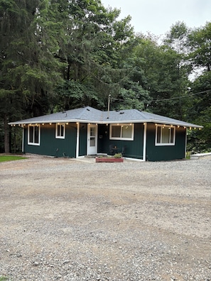 A single-story house with a gravel driveway, a front porch with a bench, and a small garden area. - House in Seaside- Elsie’s Humble Haven   Creekside view (Seaside)